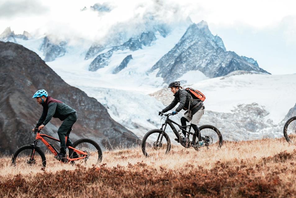 Two mountain bikers riding a trail with snow-capped Alpine peaks in the distance, during their Haute Route traverse.